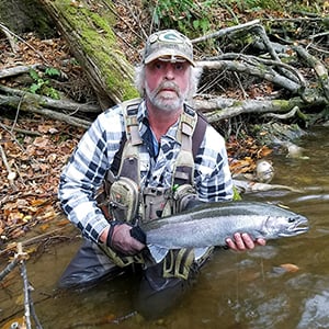 Man kneeling in stream with fishing gear and holding a caught fish