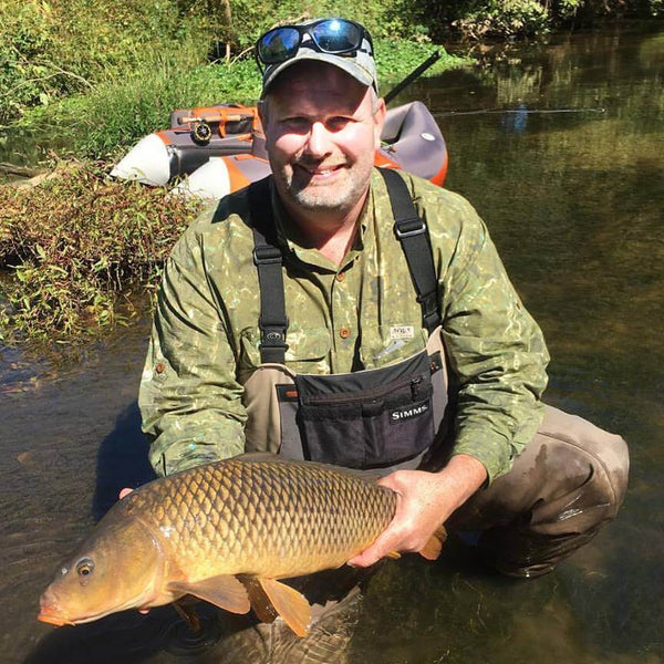 Fisherman squatting in a shallow stream while holding a brown and orange fish